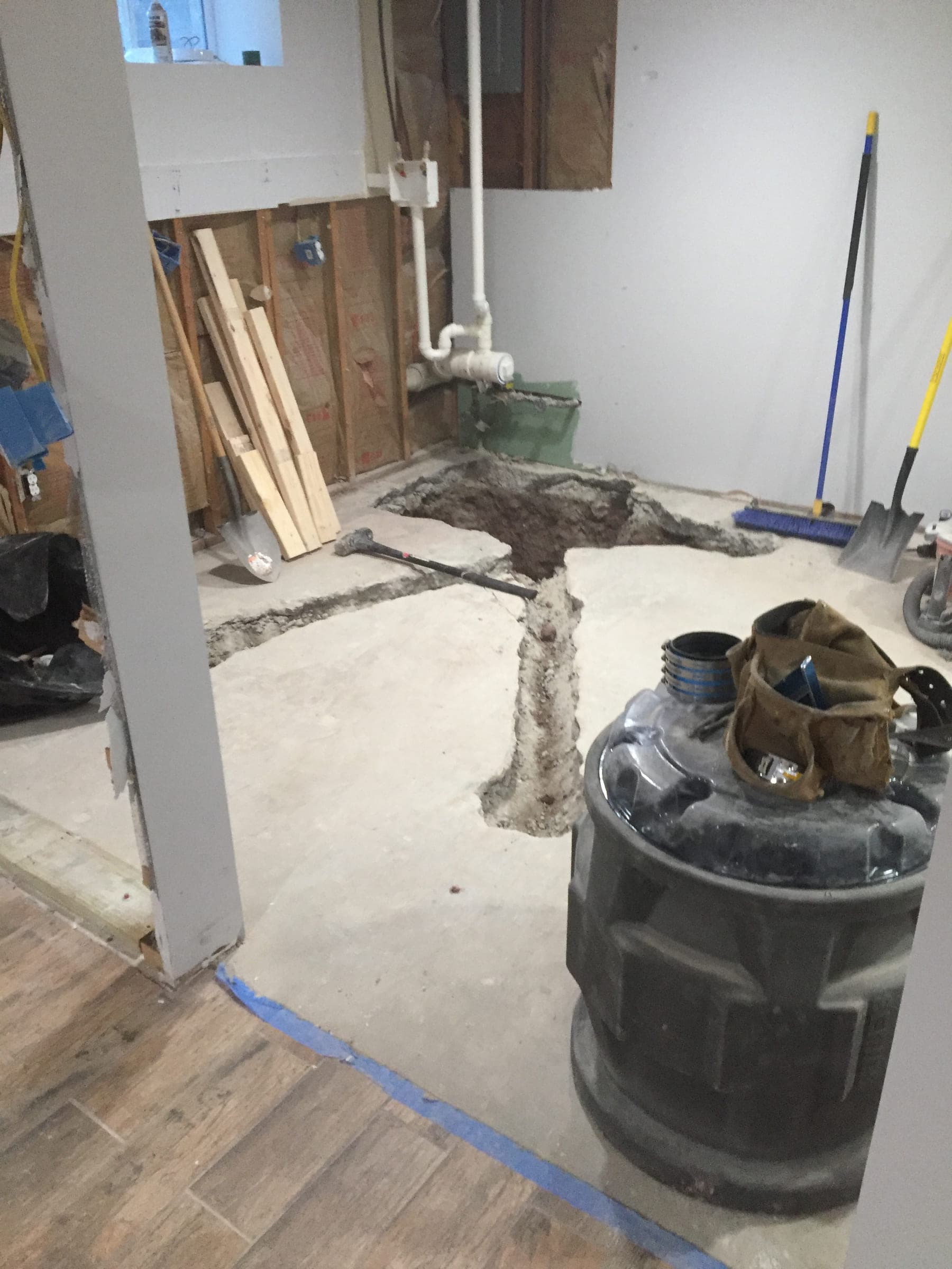 Basement floor slab cut open in a trench with a large plastic basin standing ready, beside walls stripped back to the foundation