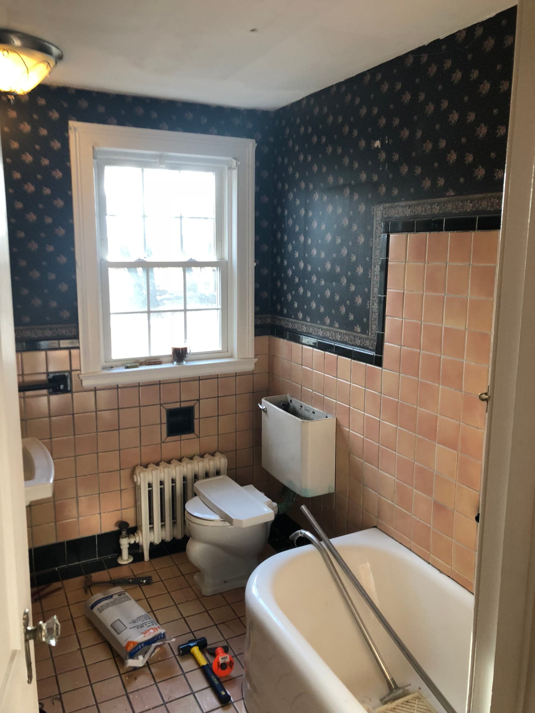 Dated bathroom before work, with pink wall tile, patterned wallpaper, an old toilet and tools on the floor