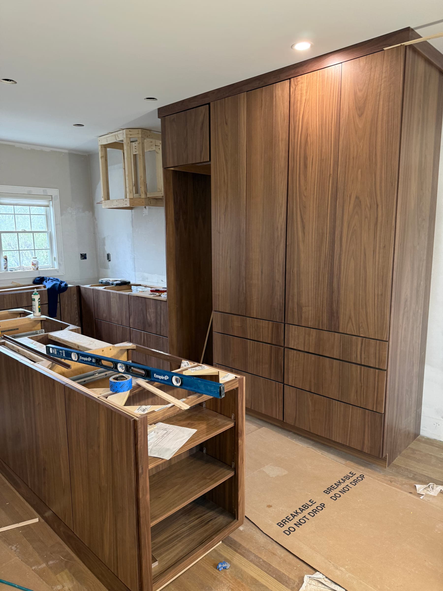 Walnut kitchen cabinetry being installed, with a run of full-height units and an island carcass levelled in place