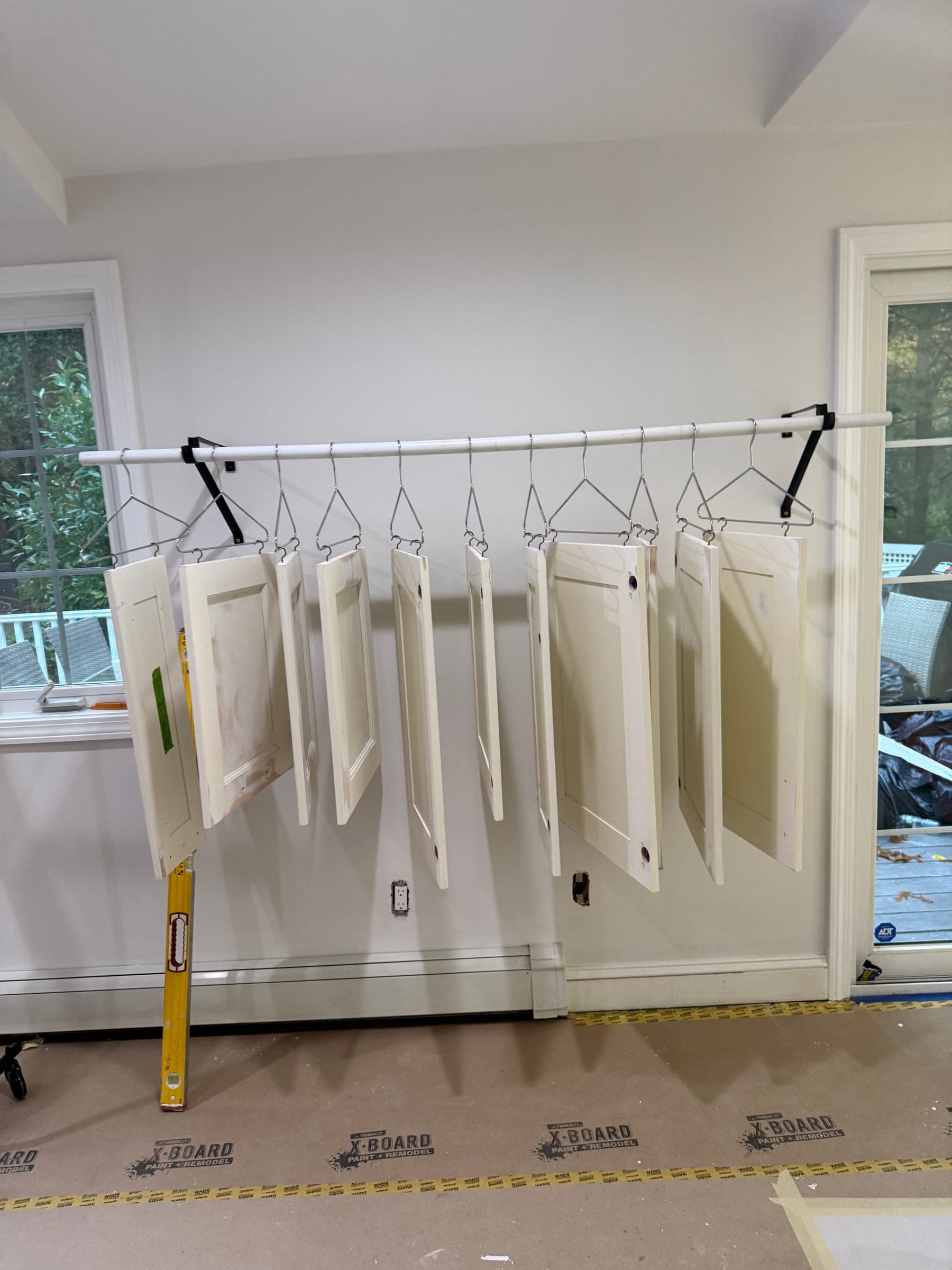 Freshly sprayed cabinet doors hanging from a mobile rack on wire hangers, above a papered and taped floor