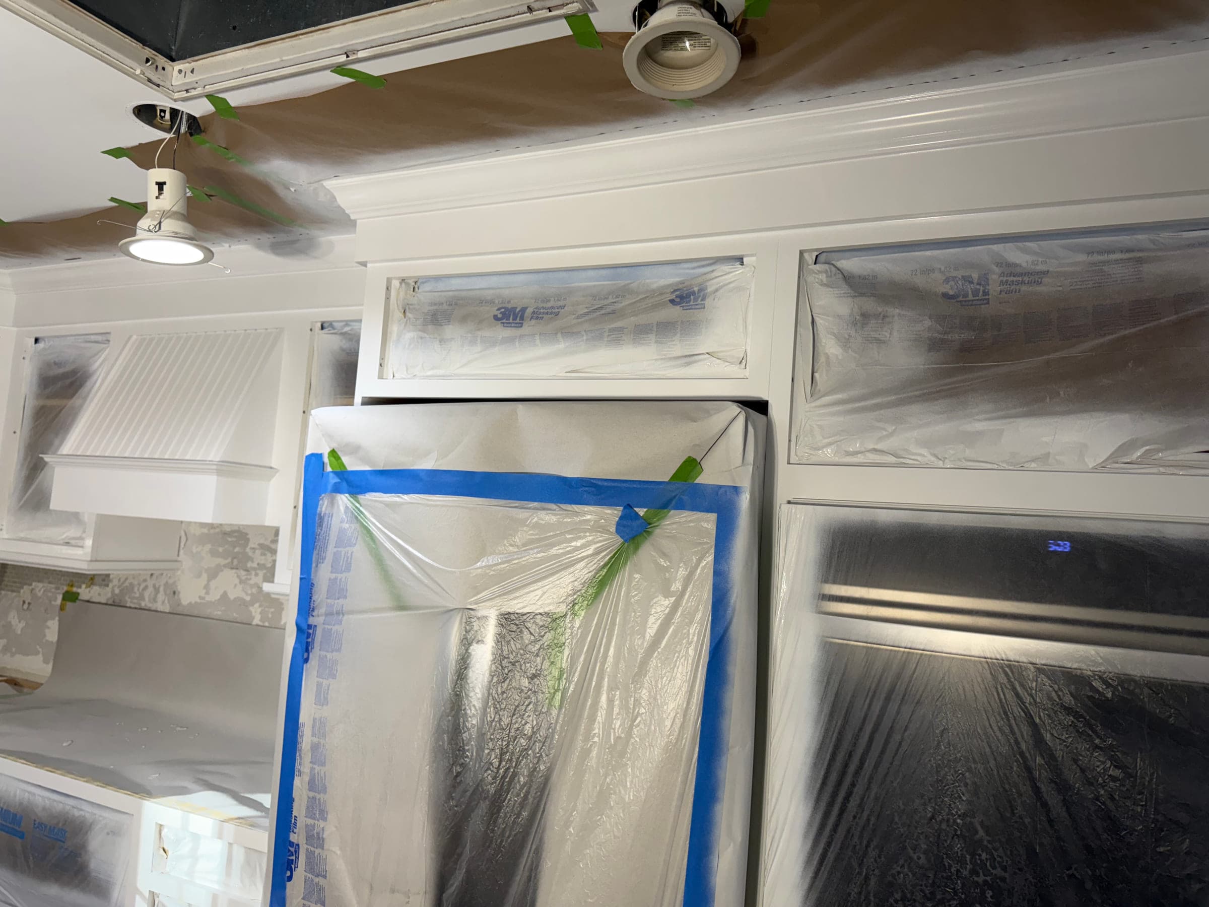 Masked kitchen wall units with taped recessed light trims and an appliance wrapped in plastic sheeting