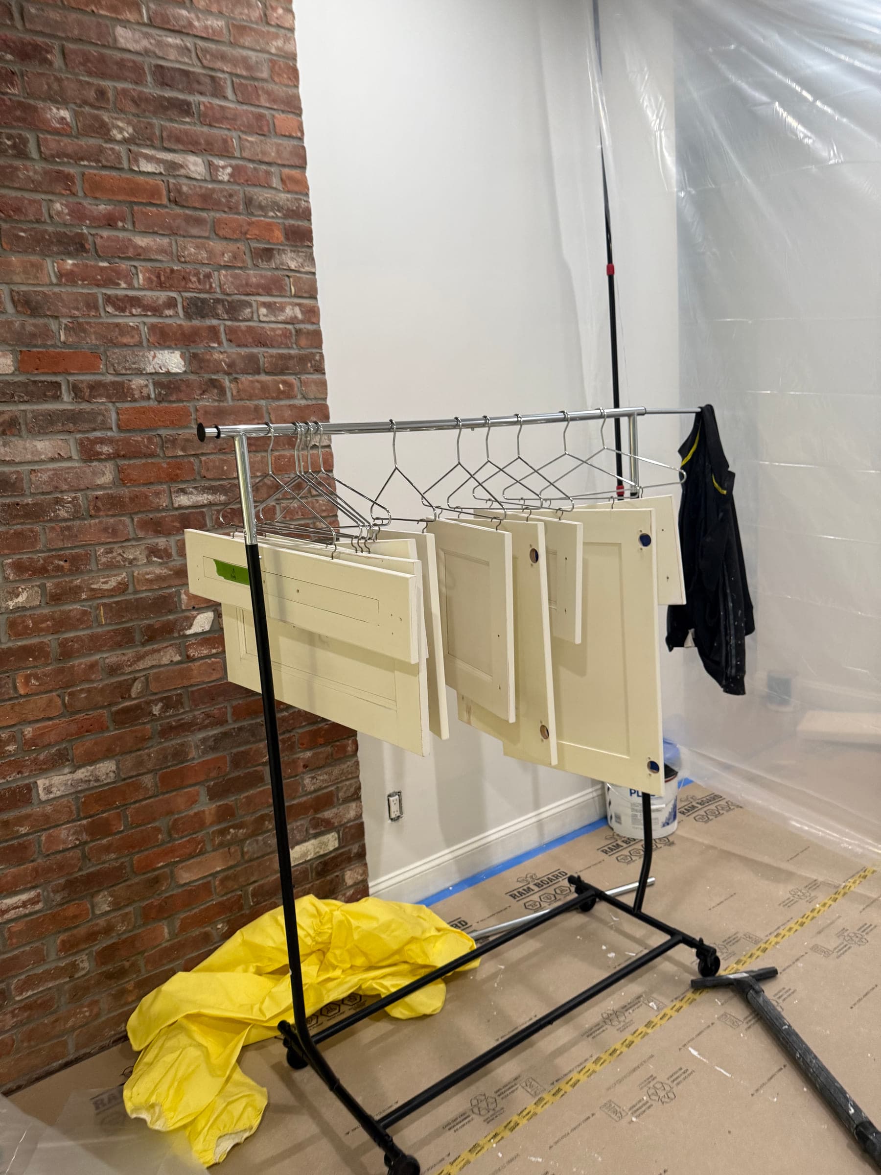 Sprayed cabinet doors racked to cure beside an exposed brick chimney breast, with sheeting on the wall and the floor protected