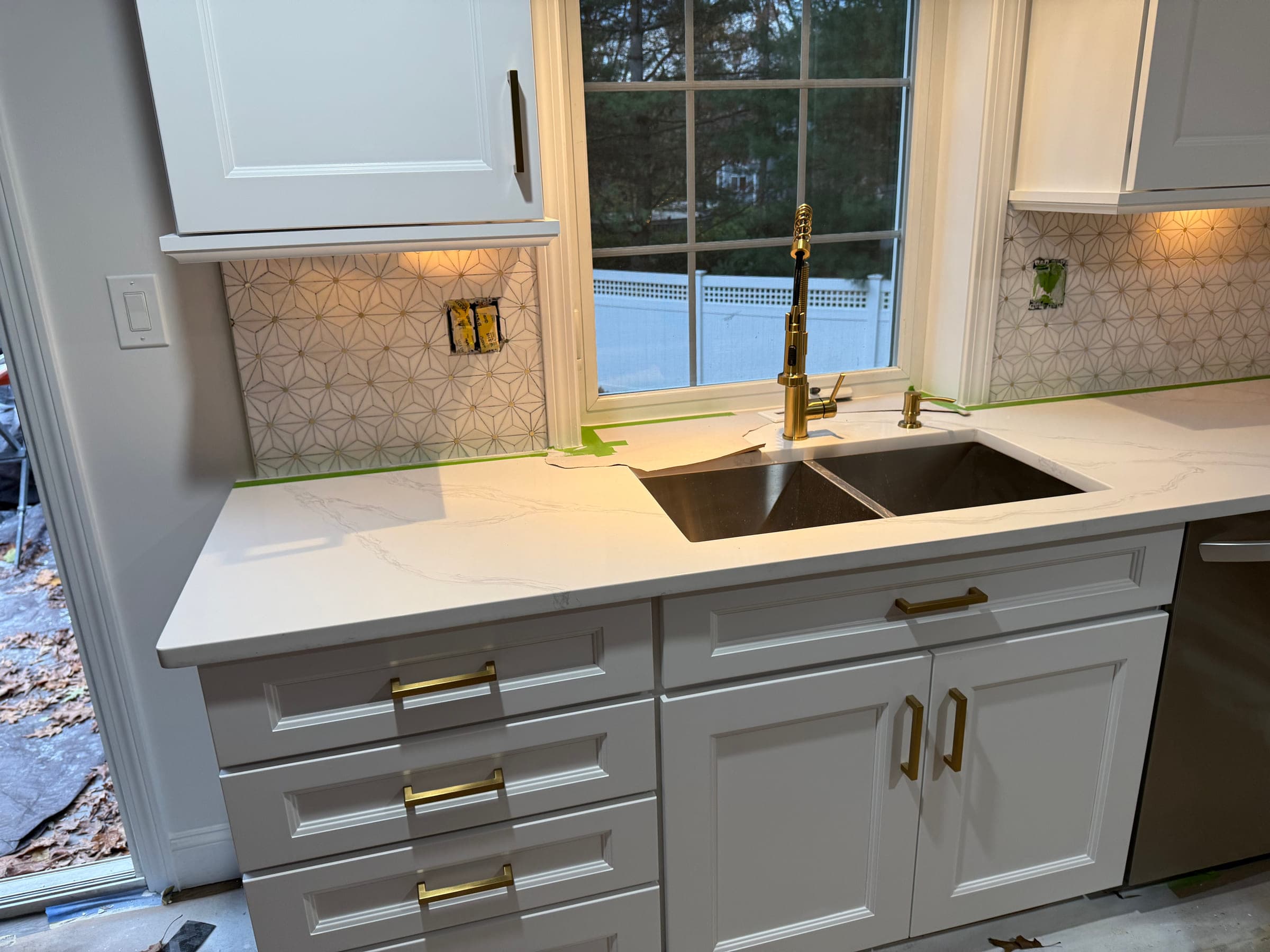 Undermount sink with a brass tap set into a quartz worktop, below a window and a patterned tile splashback